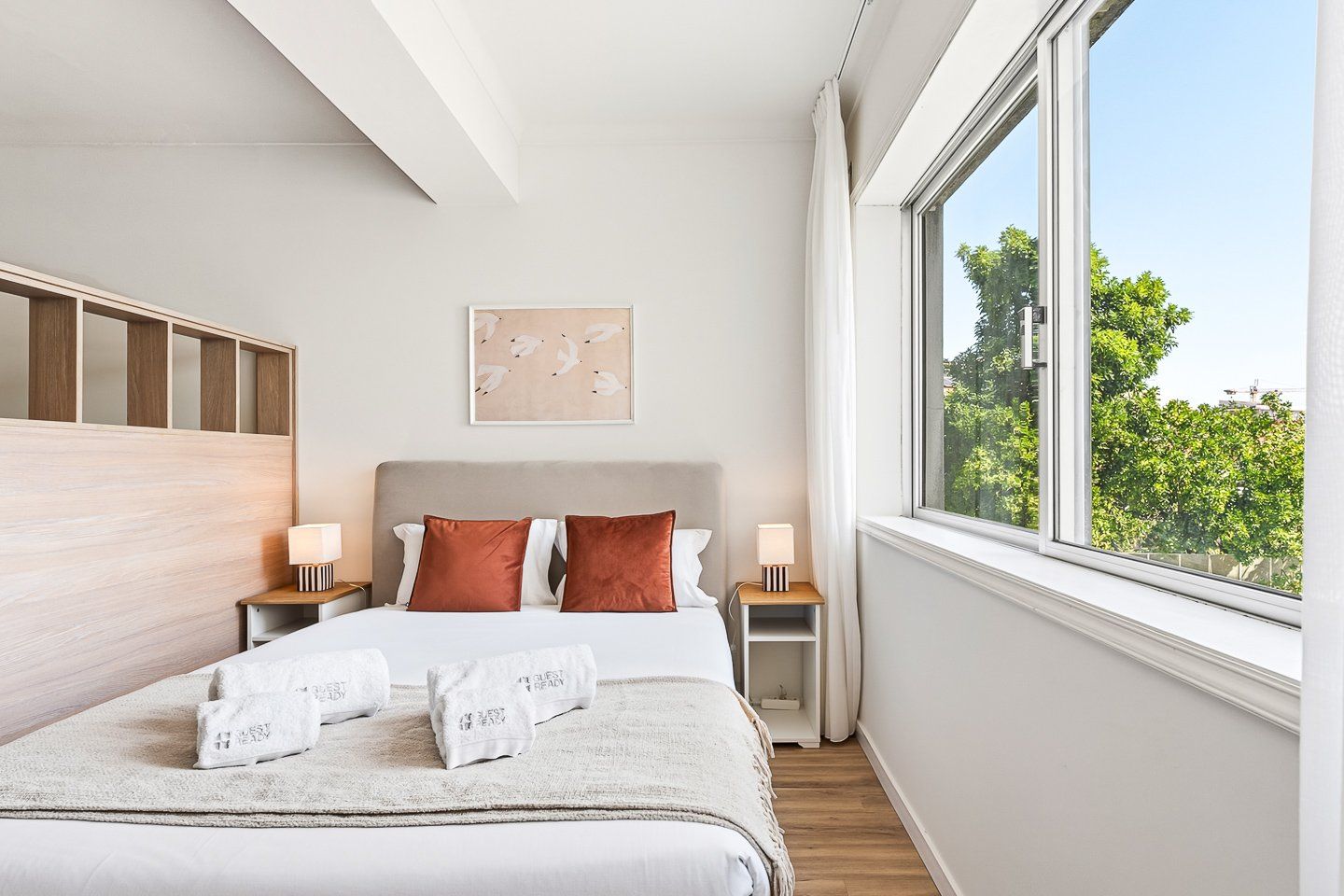 A hotel room with a bed, two nightstands, a window with a view of trees, and a wall-mounted artwork.