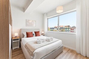 A hotel room with a large bed, a bedside table with a lamp, a window with a view of buildings, and a ceiling light. - GuestReady - Charming Studio in Porto's Heart (Porto)
