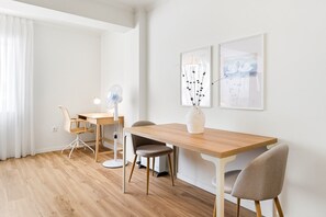 Ett modernt, minimalistiskt rum med ett matbord i trä, två stolar, en golvfläkt och två inramade konstverk på väggen.