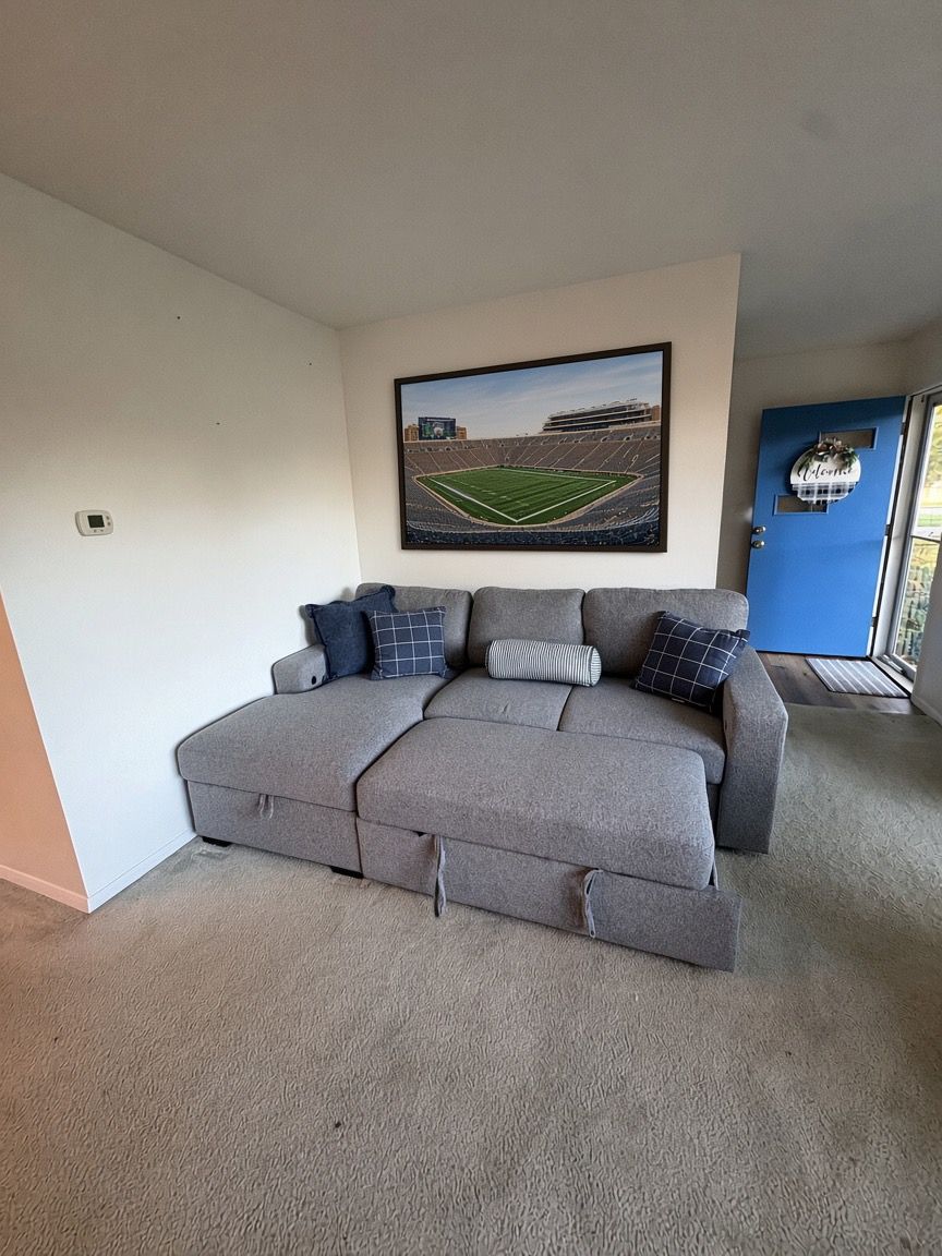 Ein graues Ecksofa mit vielen Kissen in einem Zimmer mit Teppichboden und einem gerahmten Bild an der Wand.