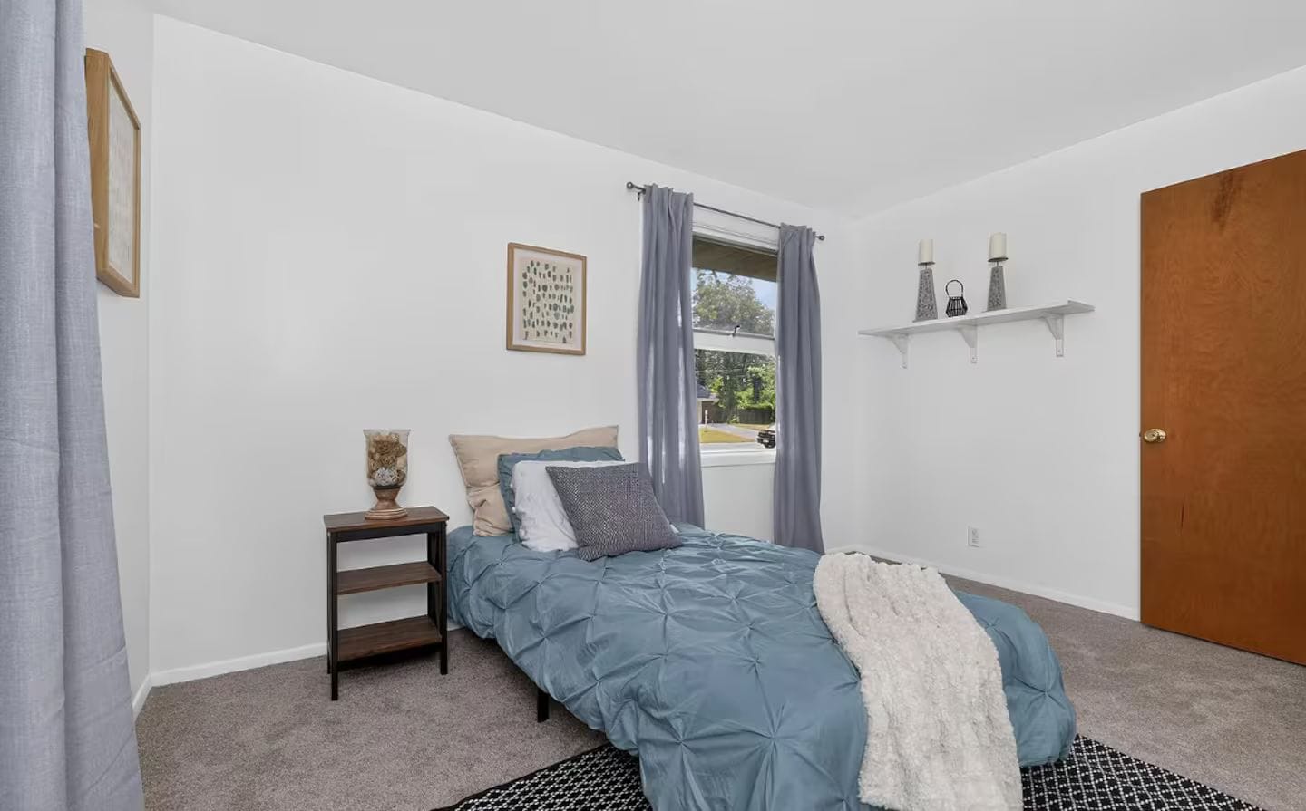 A bedroom with a bed, a nightstand, a window with curtains, a door, and a wall-mounted shelf.