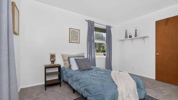 A bedroom with a bed, a nightstand, a window with curtains, a door, and a wall-mounted shelf.