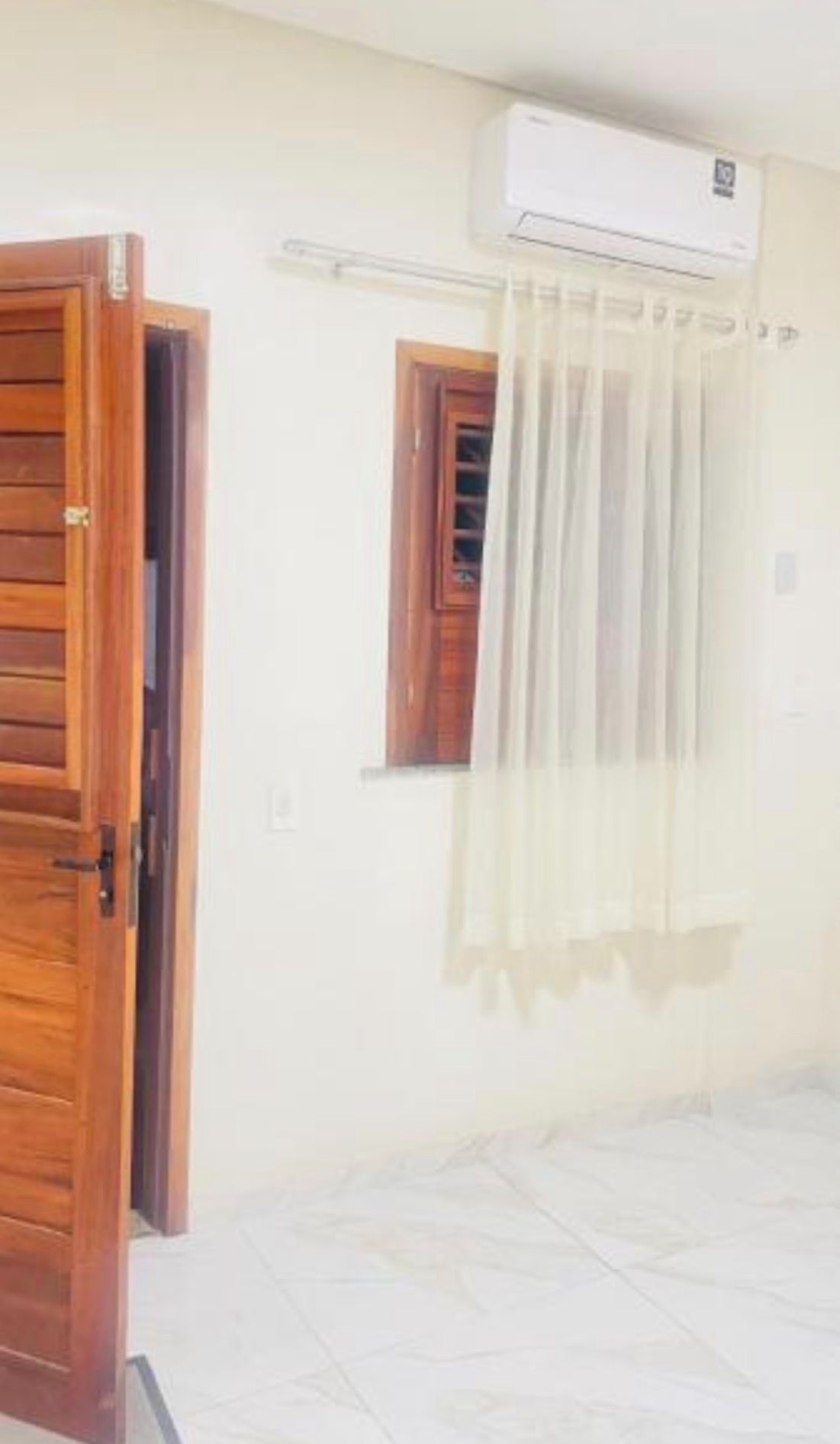A room with a wooden door, a window with a wooden frame, and an air conditioning unit mounted on the wall.