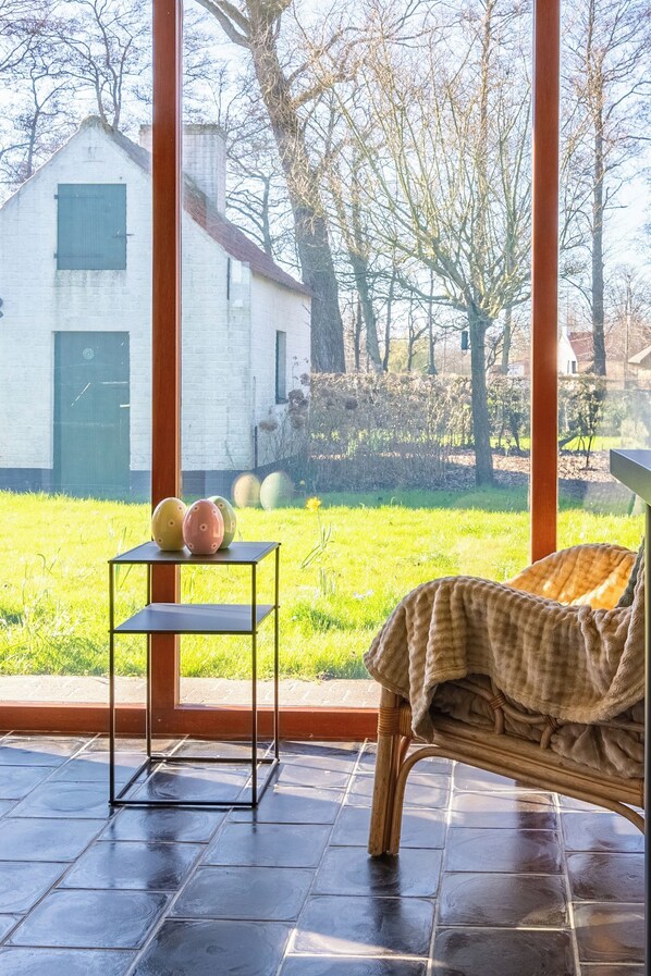 A room with a large window, a wicker sofa, a small table with decorative eggs, and a view of a white building and trees.