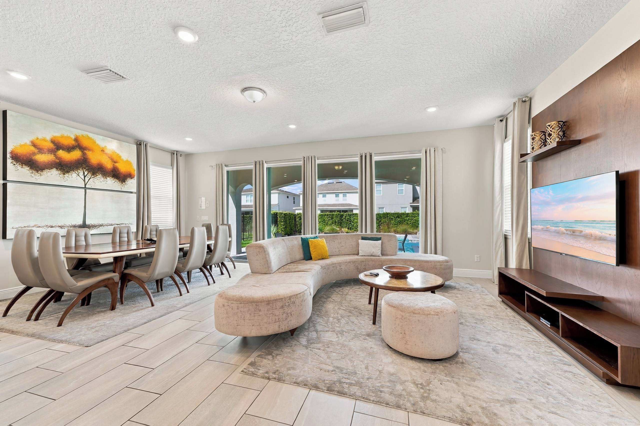 A spacious living area with a curved sectional sofa, a dining table with chairs, a flat-screen TV, and a large window overlooking a residential area.