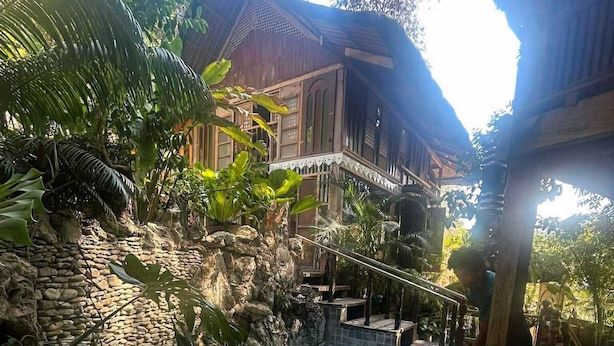Una piscina con pared de piedra y plantas tropicales, rodeada por una terraza de madera y un edificio con balcón.