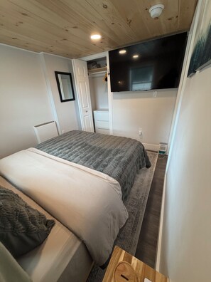 Un dormitorio con cama, mesita de noche, armario, televisor montado en la pared y una lámpara en el techo.