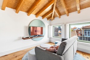 A modern living room with a television mounted on a wall, a sofa, and a window with a view of buildings. - GuestReady - Nárnia (Porto)