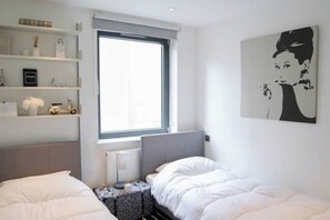 Une chambre avec deux lits, une fenêtre, un radiateur, une table de chevet et une étagère avec des objets décoratifs.