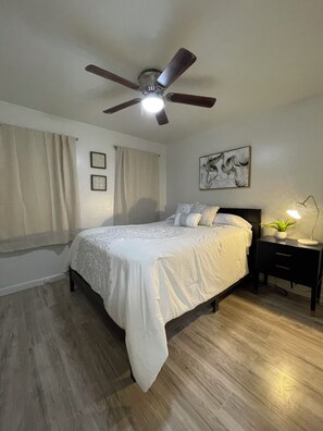 A bedroom with a bed, a nightstand, a ceiling fan, a wall-mounted lamp, and a window with curtains. - Charming 3-bedroom house in adorable Brownsville with AC (Brownsville)