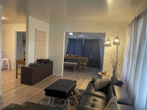 A living room with a black leather sofa, a small ottoman, a brick planter with plants, a dining area with a table and chairs, and a hallway with a staircase. - Charming 3-bedroom house in adorable Brownsville with AC (Brownsville)
