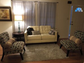 Ein Wohnzimmer mit einem beigen Sofa, gemusterten Sesseln, einem kleinen Teppich, einer Lampe und einer Tür mit Fenster.