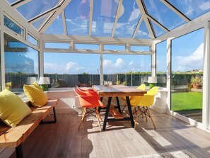 A conservatory with a wooden table and chairs, a bench, and a view of a garden. - Mermaid Gaze (Newquay)