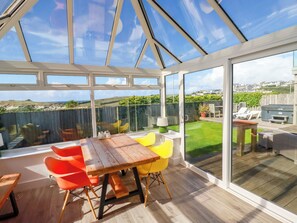 A conservatory with a wooden table and chairs, a view of the garden, and a patio area with outdoor furniture. - Mermaid Gaze (Newquay)