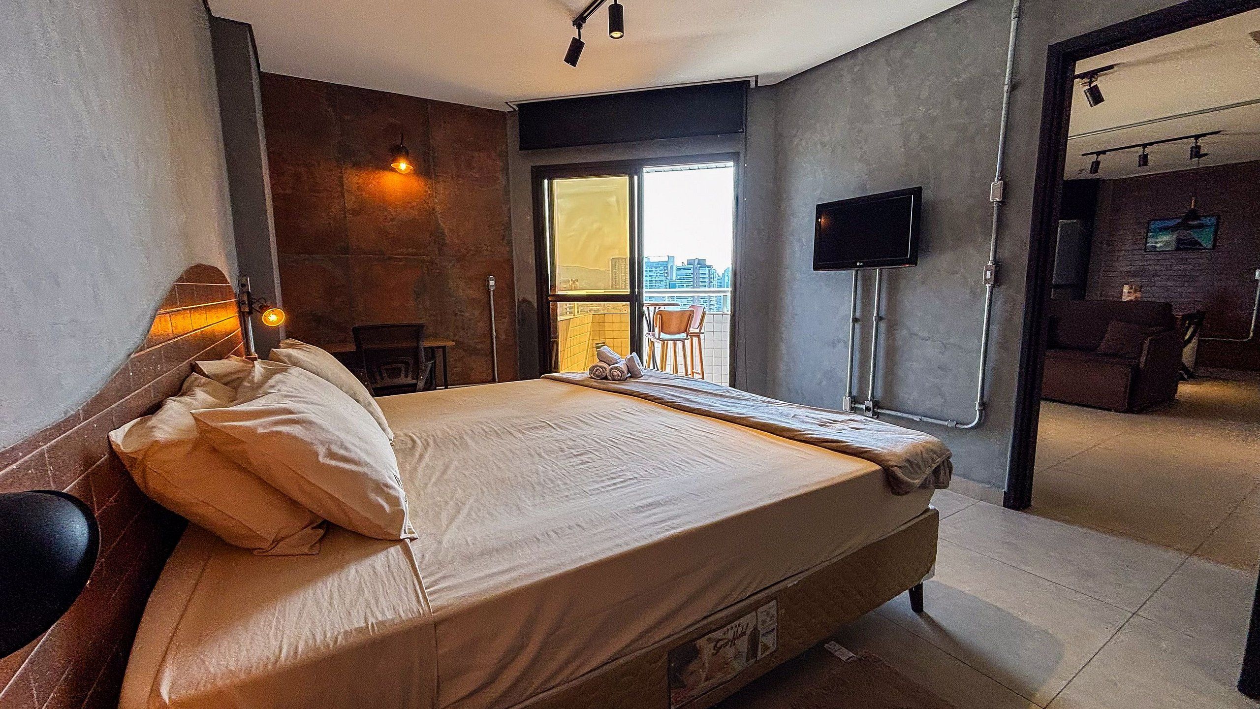 A hotel room with a bed, a TV, a balcony with a chair, and a view of the city.