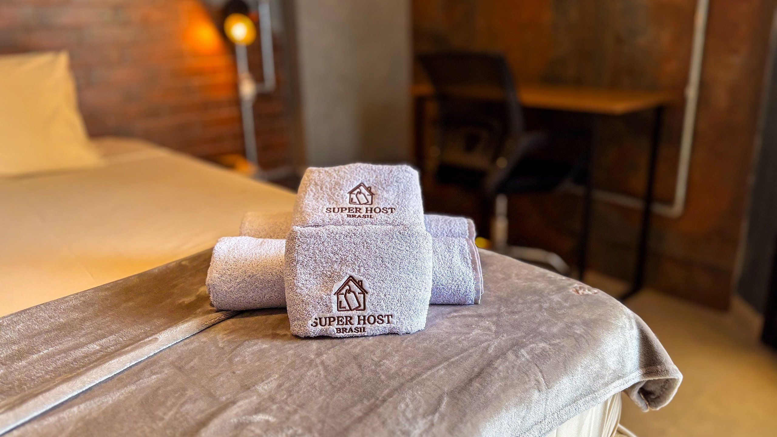 A bed with a grey textured blanket and two folded towels with 'SUPER HOST BRASIL' branding.