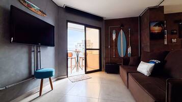 A modern living room with a flat-screen TV, a sofa, a coffee table, and a view of the city through a glass door.
