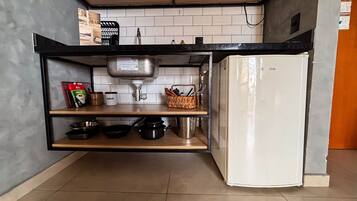 Cocina compacta con un refrigerador blanco, una encimera negra y una estantería con utensilios de cocina y una botella.
