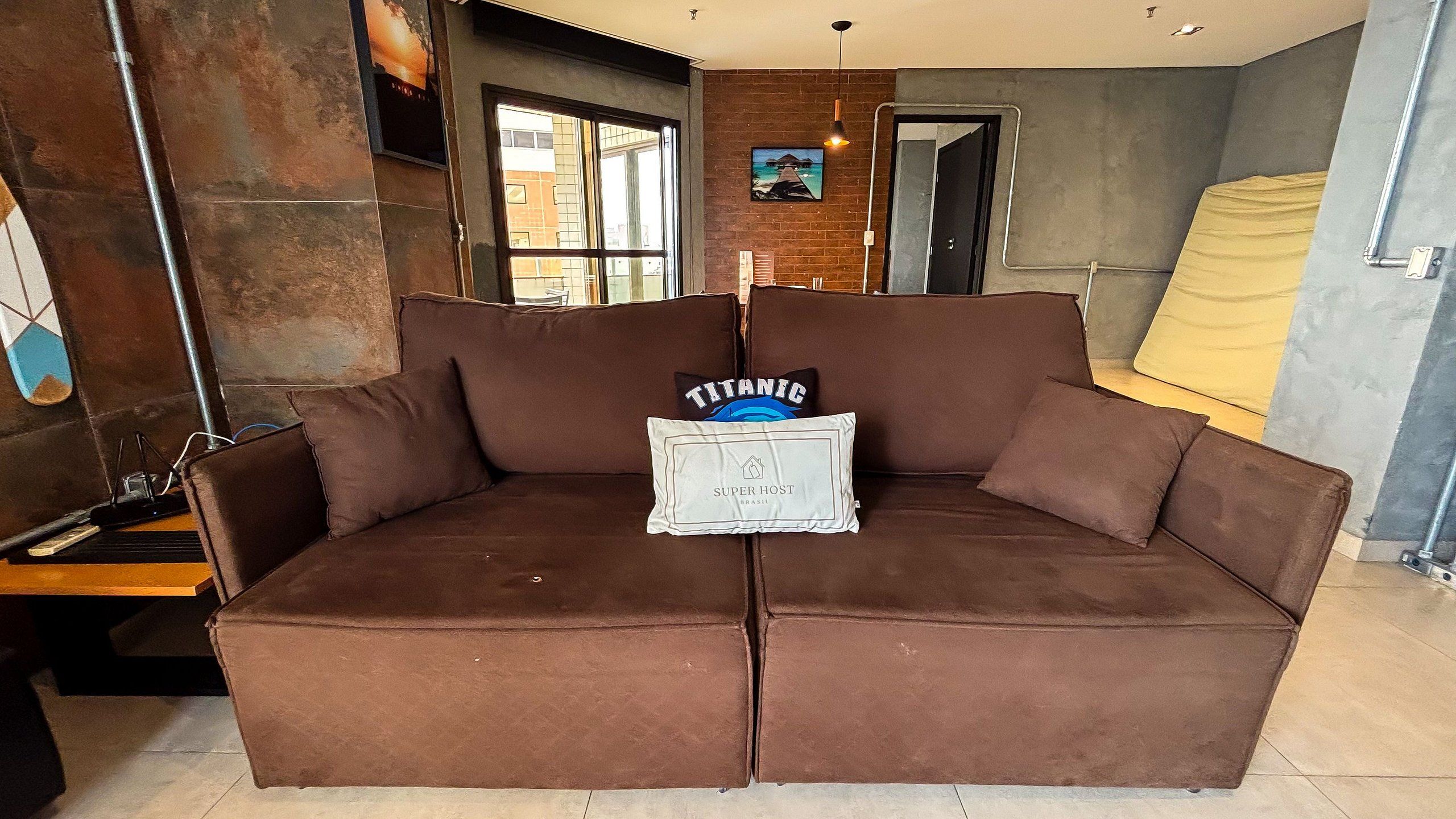 A brown sofa with a pillow labeled 'TITANIC' and 'SUPER HOST BRASIL' in a room with a brick wall and a window.
