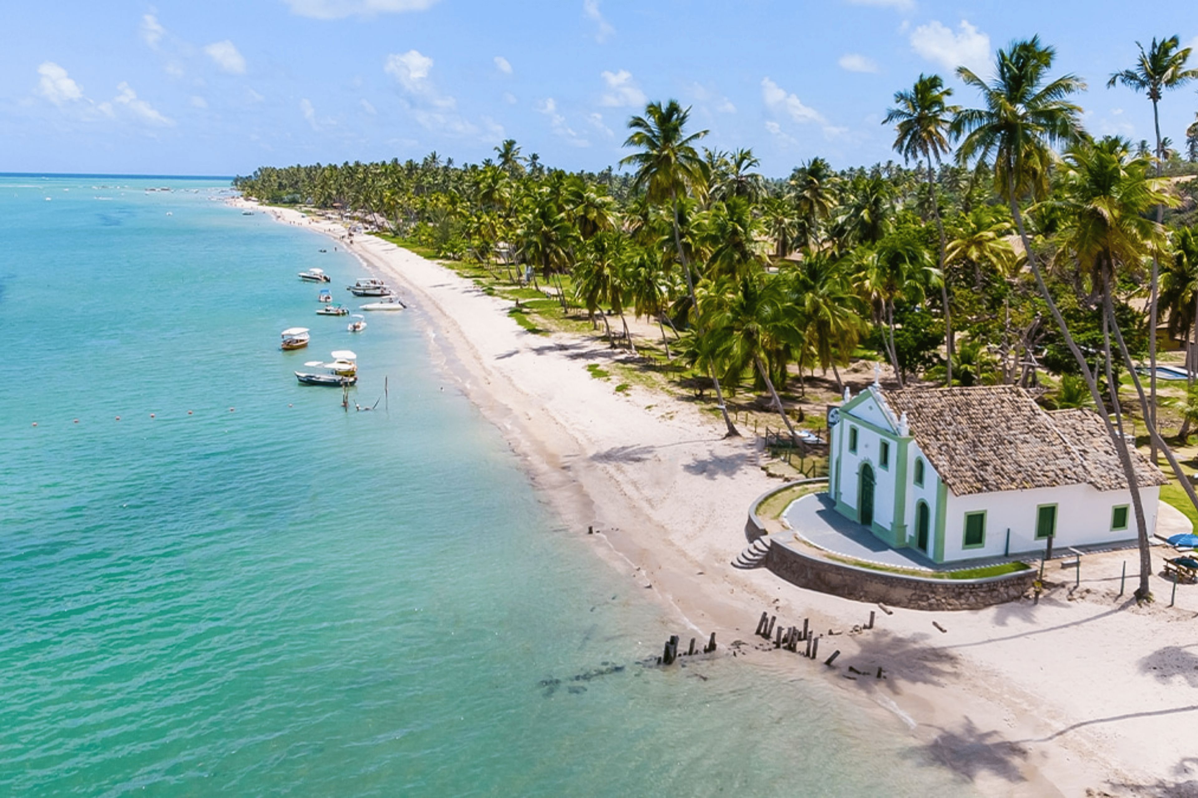 A beach with clear turquoise waters, a row of palm trees, and a small white building with a thatched roof.