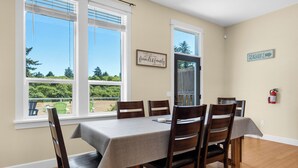 Un comedor con una mesa dispuesta para seis personas, sillas de madera, ventanas amplias con persianas y un letrero enmarcado en la pared. También se observa un extintor colgado en la pared.