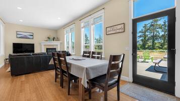 Un coin repas avec une table dressée pour six personnes, flanqué de deux fenêtres équipées de stores, d’une cheminée et d’un téléviseur en arrière-plan.