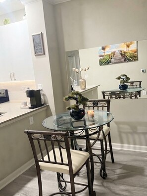 Un comedor con una mesa redonda de vidrio, cuatro sillas, un florero con flores y un cuadro en la pared.