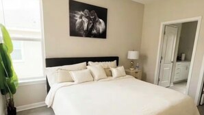 A bedroom with a large bed, a ceiling fan, a window with curtains, a wall-mounted picture, and a door leading to another room. - King Suite In Charlotte  (Charlotte)
