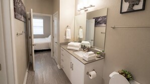 A bathroom with a toilet, sink, and mirror; a towel rack with towels; a framed picture on the wall; and a door leading to a bedroom. - King Suite In Charlotte  (Charlotte)