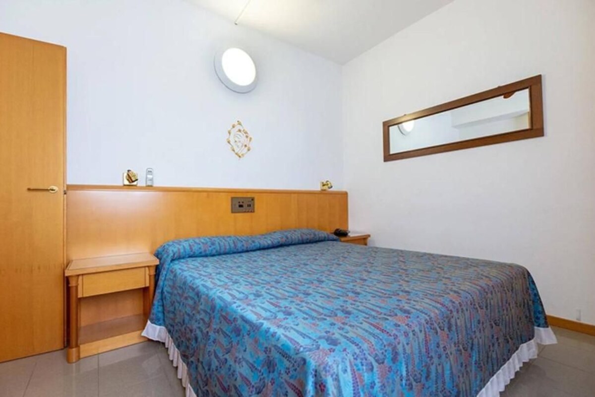 A bedroom with a bed, a bedside table, a nightstand, a wall-mounted mirror, and a door.