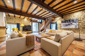 A spacious living room with a stone wall, wooden beams, a large sofa set, a television, and a staircase leading to an upper level. - La Guate - Maison avec piscine - Saint-Émilion (Daubèze)