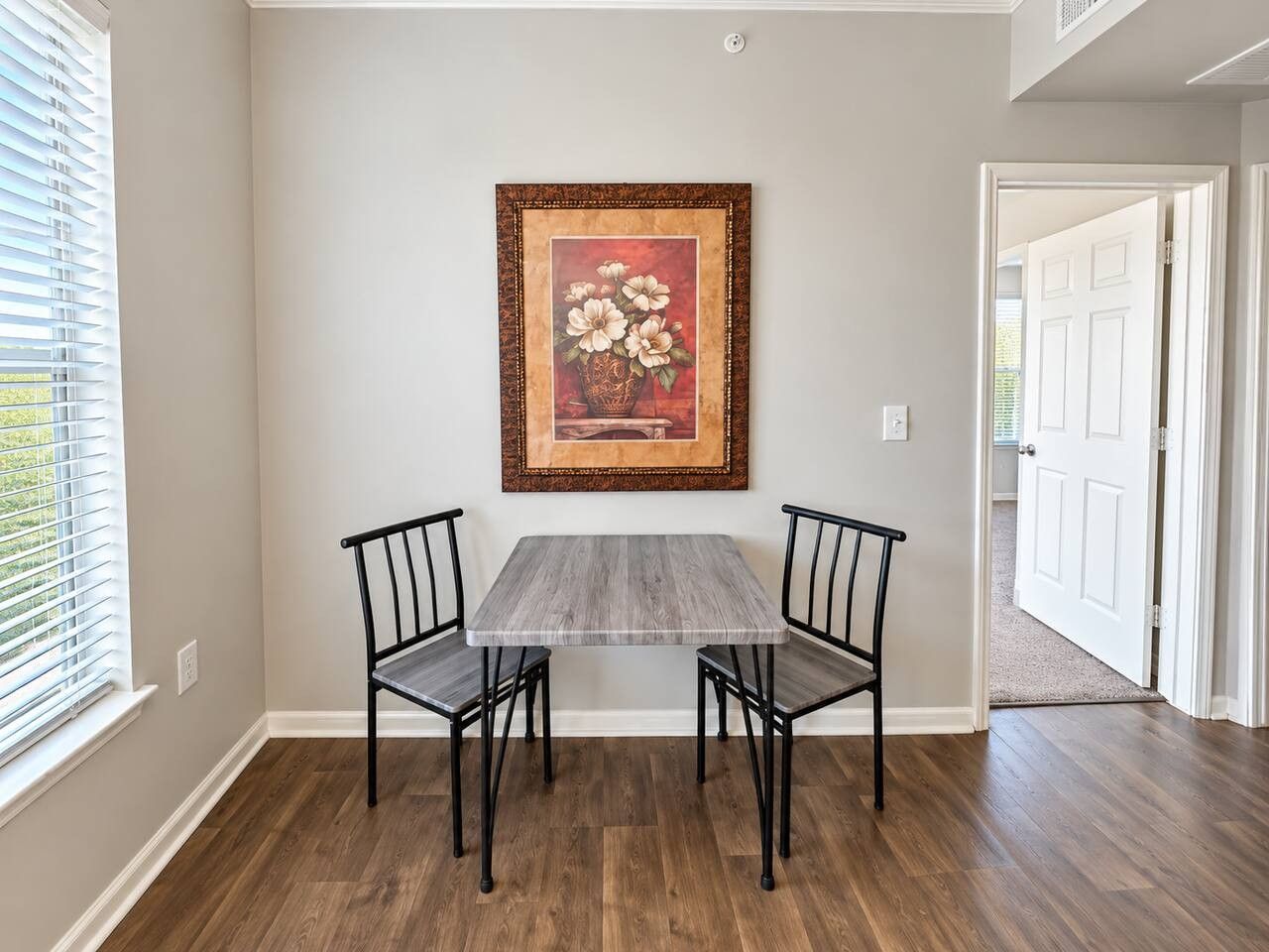 Un coin repas avec une table et des chaises, un tableau encadré au mur et une porte donnant sur une autre pièce.