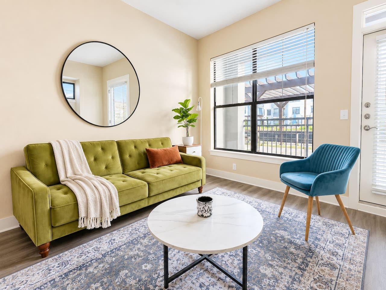 Un salon avec un canapé vert, un fauteuil bleu, une table basse ronde et une grande fenêtre avec des stores.