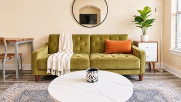 Un salon avec un canapé vert, une table basse ronde, un téléviseur fixé au mur, une plante en pot et un lampadaire.