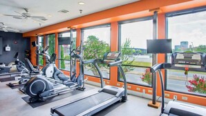 Une salle de sport équipée de tapis de course, de vélos elliptiques et de vélos d’appartement, de grandes fenêtres offrant une vue sur la ville et de ventilateurs de plafond.