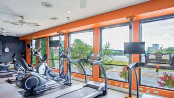 Ein Fitnessstudio mit Laufbändern, Crosstrainern und Fahrrädern, großen Fenstern mit Blick auf die Stadt und Deckenventilatoren.