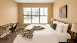 Une chambre d’hôtel avec un lit, un bureau, une chaise, un ventilateur de plafond, une fenêtre avec des stores et un tableau au mur.