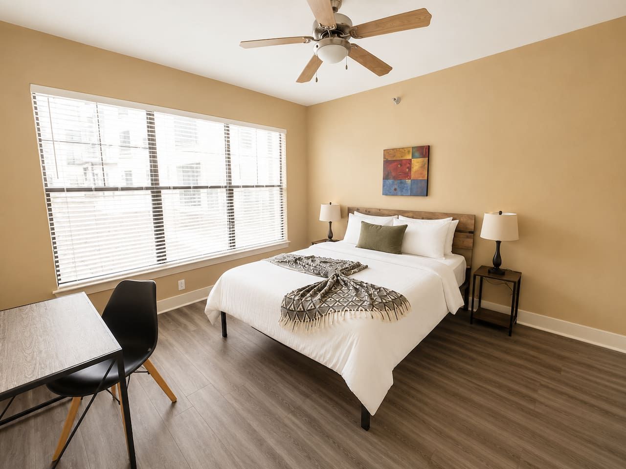 Une chambre d’hôtel comprenant un lit, deux tables de chevet avec des lampes, un bureau avec une chaise et une fenêtre avec des stores.
