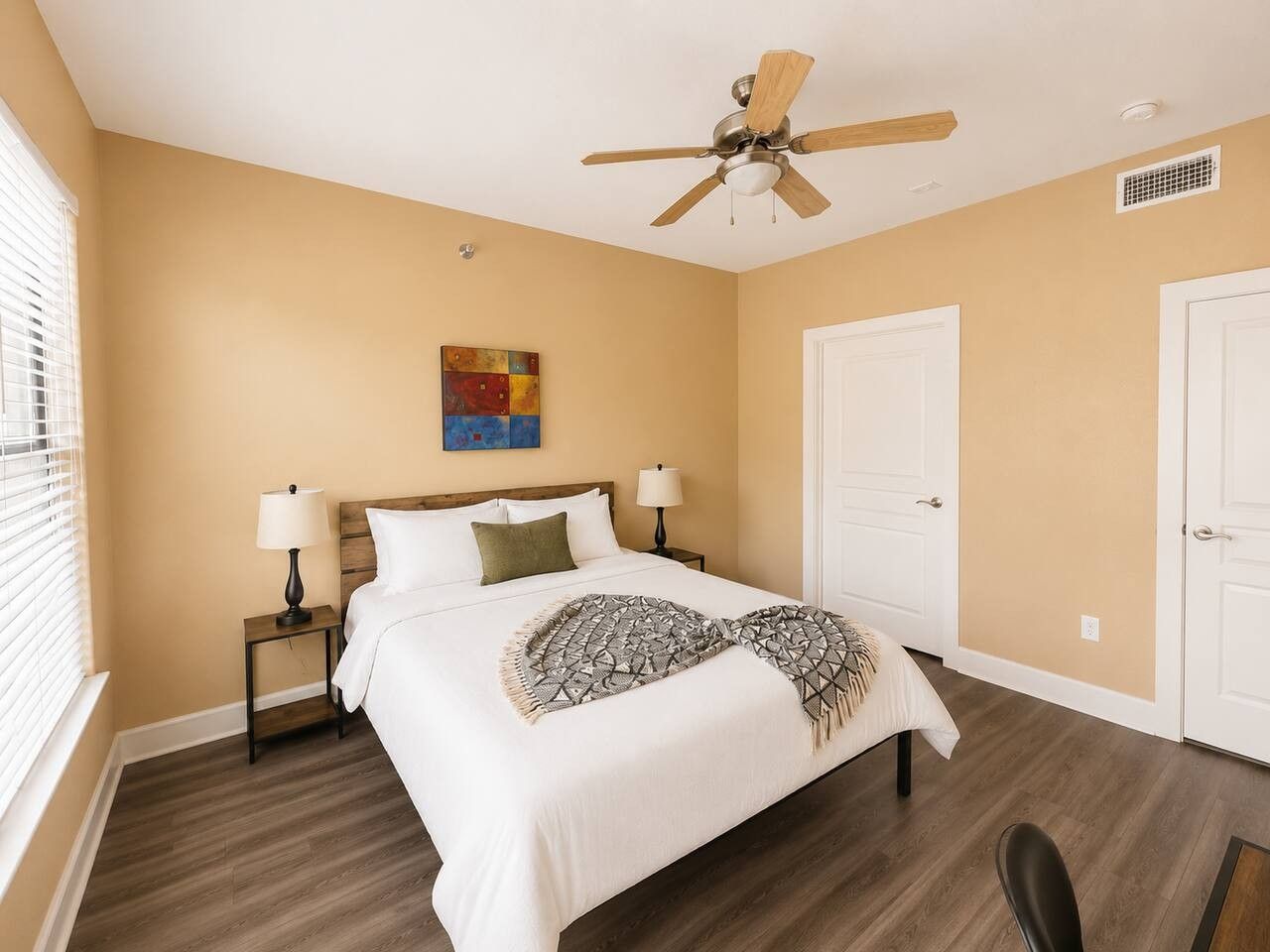 A bedroom with a bed, two nightstands, a ceiling fan, a door, and a window with blinds.