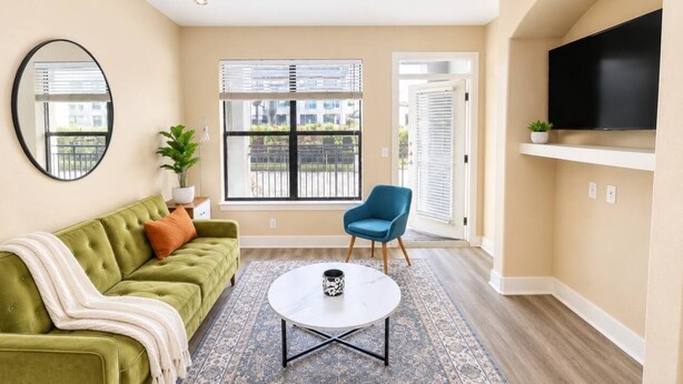 Un salon avec un canapé vert, un fauteuil bleu, une table basse ronde et un téléviseur fixé au mur.