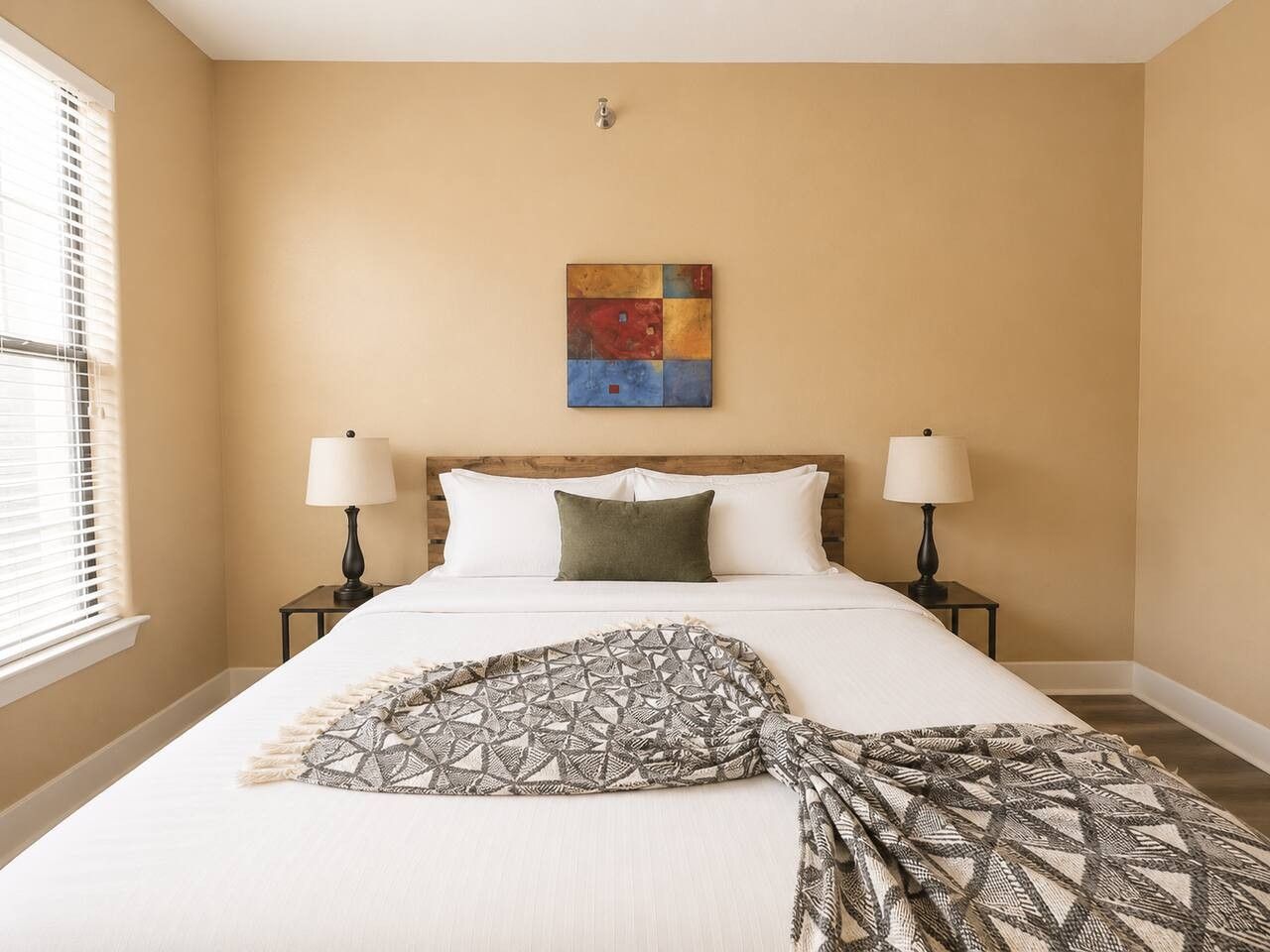 A hotel room with a bed, two bedside lamps, a window with blinds, and a wall-mounted artwork.
