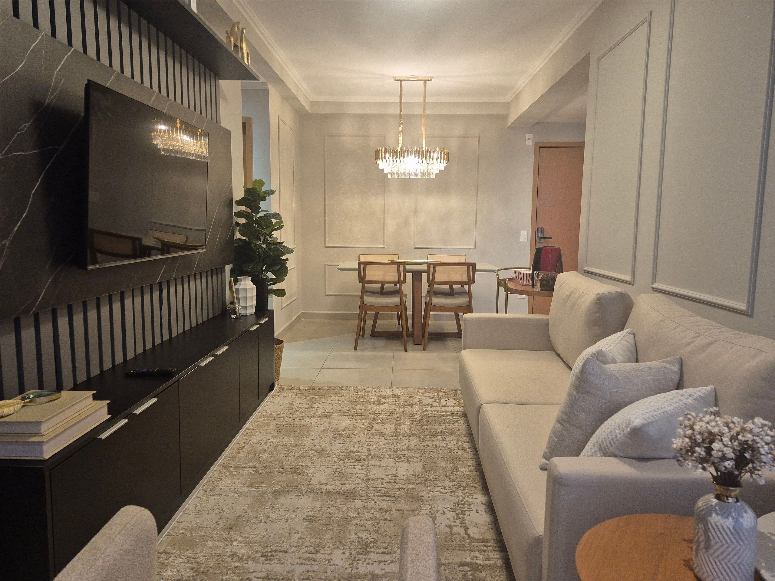 A modern living room with a flat-screen TV, a dining area with a chandelier, and a sofa with decorative pillows.