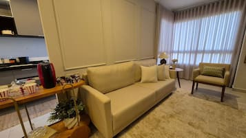 Un salon comprenant un canapé, des fauteuils, une table basse, une lampe, et un coin cuisine à l’arrière-plan.