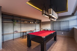 Uma mesa de bilhar moderna em ambiente interno, com superfície de feltro vermelho, rodeada por piso de madeira e prateleiras com diversos objetos.