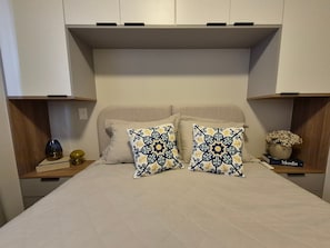 A bedroom with a bed, two pillows, a bedside table with a vase, and a book titled 'Moda'. - PP - Neo Petrópolis - Apto NP148 (Presidente Prudente)