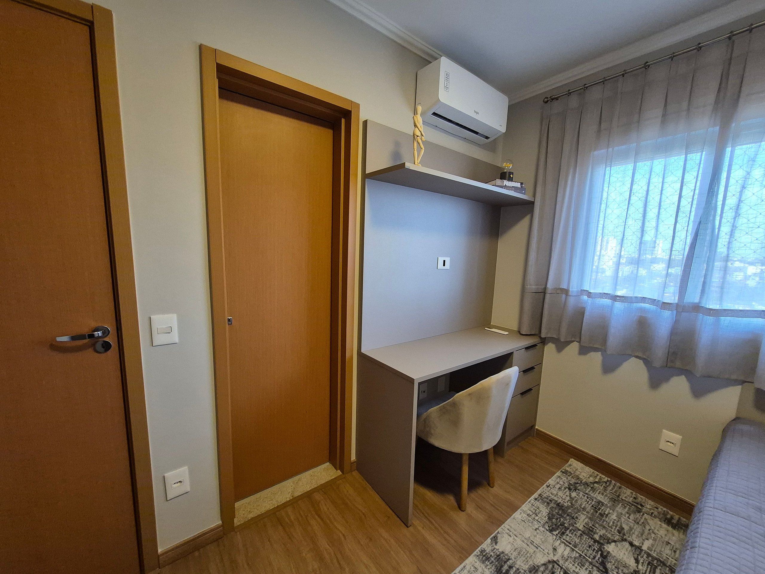 A room with a wooden door, a desk with a chair, an air conditioning unit, and a window with curtains.