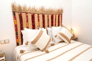 Cama com roupa de cama branca e almofadas decorativas, cabeceira com padrão trançado e luminária de cabeceira.