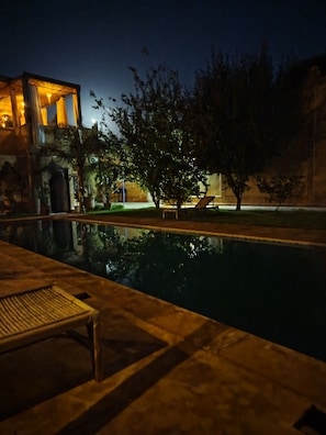 Vista noturna de uma área de piscina com espreguiçadeira e prédio com janelas iluminadas.