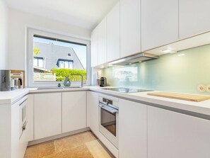A modern kitchen with white cabinetry, a built-in oven, and a window overlooking a house. - Holiday Home Heide (Sylt)
