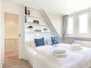 Ein Schlafzimmer mit einem Bett, Kissen, einem Regal mit Büchern und Dekorationsobjekten, einem Fenster mit Vorhängen und einer Tür zu einem anderen Raum.